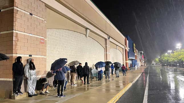 A long line of people stand at night in the rain outside of a Washington DC Best Buy to get PS5 and Xbox Series X/S