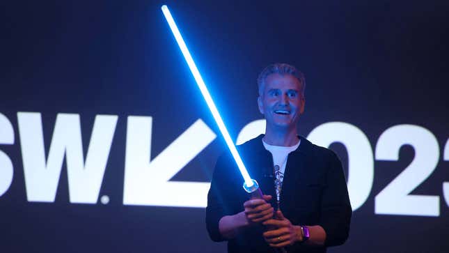 A man in a black jacket holds a glowing blue lightsaber. 
