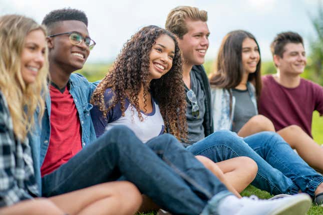 A small group of teenagers sit in the grass as they hang out talking and enjoying a few laughs together. They are each dressed casually and are smiling as they enjoy the fresh air.