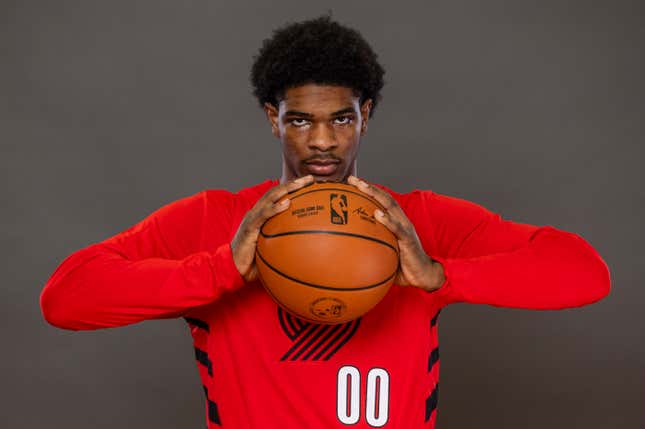 A young Black man with curly hair, wearing a red long-sleeved Portland Trailblazers shirt, holds a basketball between both hands and stares into the camera.