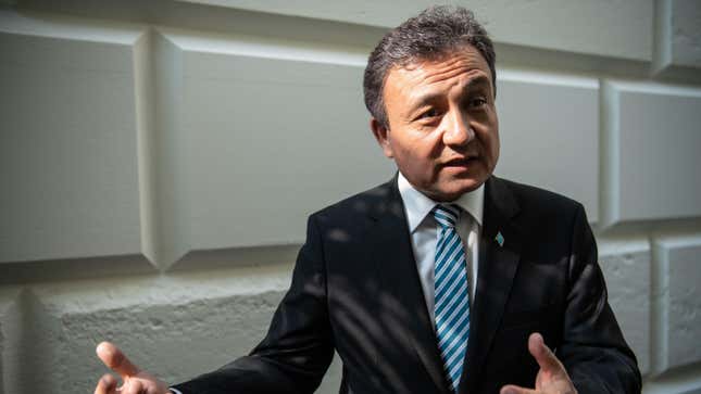 Dolkun Isa stands with his back to a white brick wall, gesturing to his right with his hands.