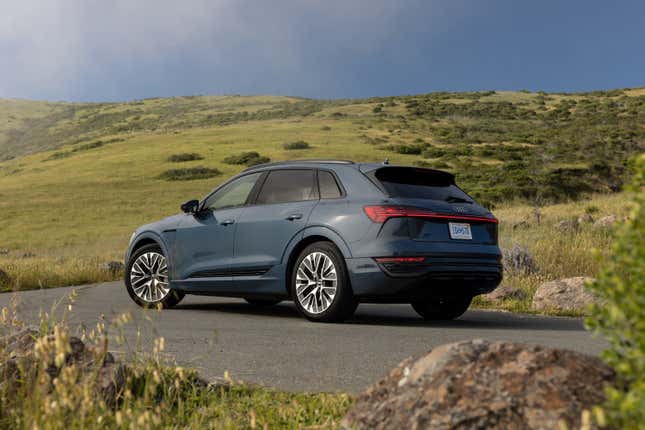 A gray-blue 2024 Audi Q8 E-Tron SUV is parked in front of green hills. Rear three-quarter view.