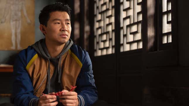 Simu Liu sits while wearing a blue and yellow jacket over a grey sweatshirt in Shang-Chi and the Legend of the Ten Rings. He's holding what appears to be a red dragon figure.