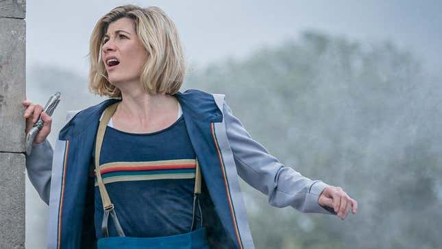 Jodie Whittaker's 13th Doctor rests against a stone column with her sonic screwdriver in hand, glancing at an unseen threat in the sky.