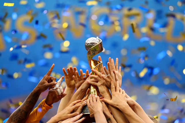 Hands are visible lifting a golden football trophy after victory in the 2019 FIFA Women's World Cup. Gold and blue confetti falls in the background.