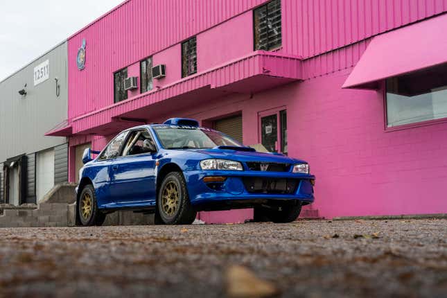 a blue vintage subaru 2.5rs coupe parked in front of a bright pink building on an overcast day. the subaru is built for rally racing, with oversized all-terrain tires and fender flares and scoops. a roll cage can be seen through the car's windows