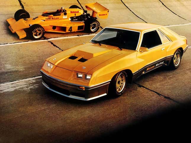 A papaya orange M81 Mustang sits on a banked race track next to an open wheel race car in the same shade.