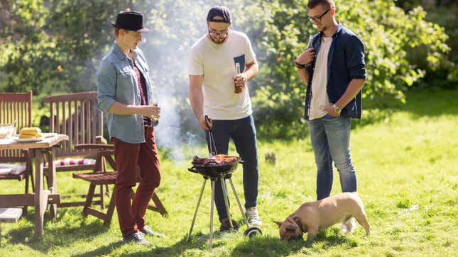 两个年轻的白人的照片在休闲装看第三个小圆木炭烤架烤香肠。有一个小的浅棕色的斗牛犬站右边的照片。