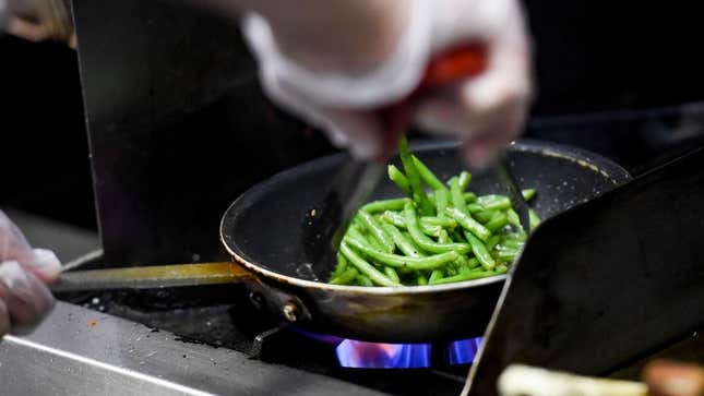A cook in a restaurant kitchen sauteing green beans in a pan on the stove