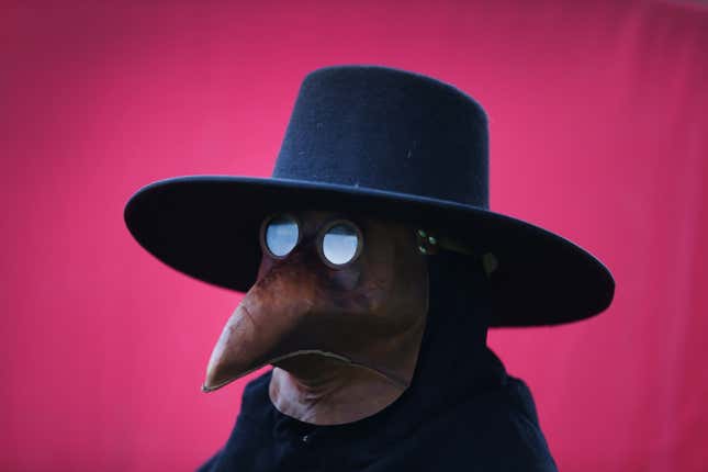 A person is dressed as a plague doctor against a bright pink backdrop. 