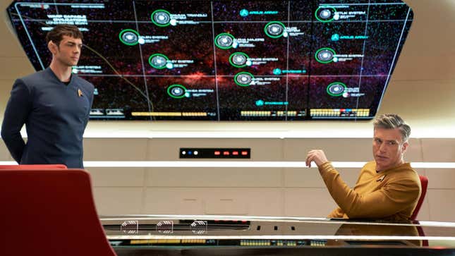 Ethan Peck's Spock (left) stands at attention in front of a large star chart, while Anson Mount's Captain Pike (right) sits in a seat, looking away from the map with his arm raised.