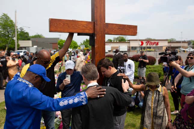 Several people stand around a giant cross marking a memorial for the people who died at the Buffalo shooting in May. They have their heads bent as they pray.
