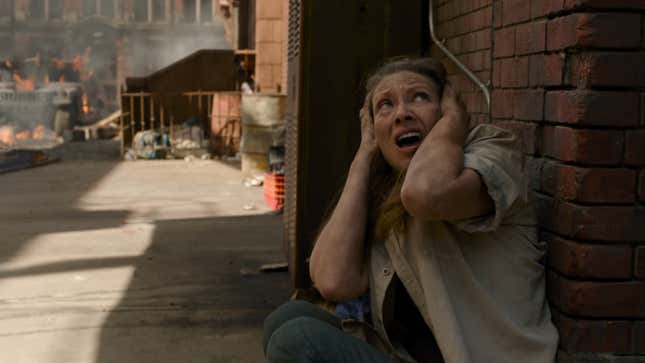 Tess is seen crouched down next to a brick building covering her ears with a frightened expression.