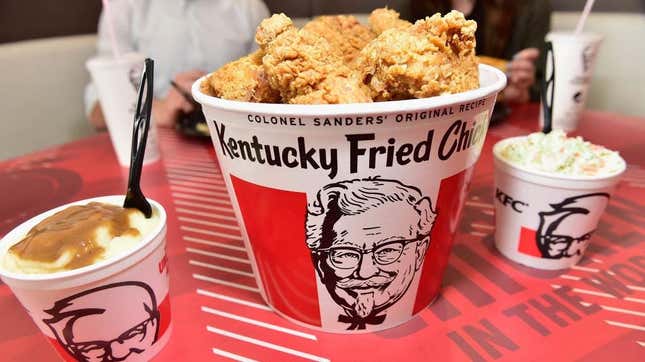 Bucket of KFC chicken, KFC mashed potatoes and gravy, and KFC cole slaw on red tabletop