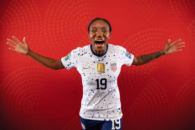 A Black woman in a white jersey and blue shorts bearing the number 19 holds our her arms and yells towards the camera in a light-hearted manner