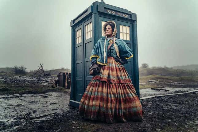 A woman in a bonnet, a colonial blue jacket, and a long red dress stands in front of the TARDIS.