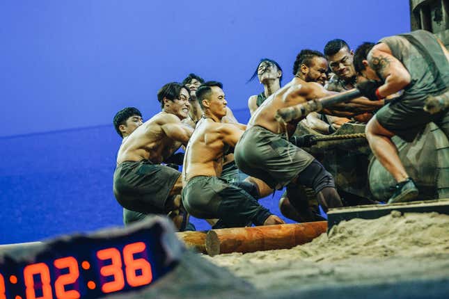 A still image shows Physical 100 contestants pulling a 1.5-ton boat across sand.