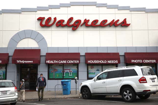 Pedestrians walk towards a Walgreens store entrance. Two cars are parked outside.