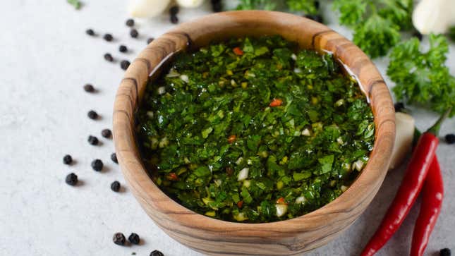 A bowl of vibrantly green chimichurri sauce in a wood bowl on a white marble counter with chili peppers, parsley, and peppercorns