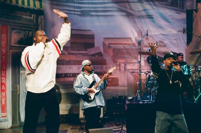 Two Black men, one tall and wearing a white sweatshirt and another smaller, wearing a black cap and sweater, rap into microphones, while two more Black man behind them play guitar and drums.