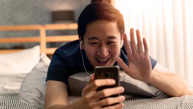 An Asian man is seen using a smartphone to have a video call while lying on a bed