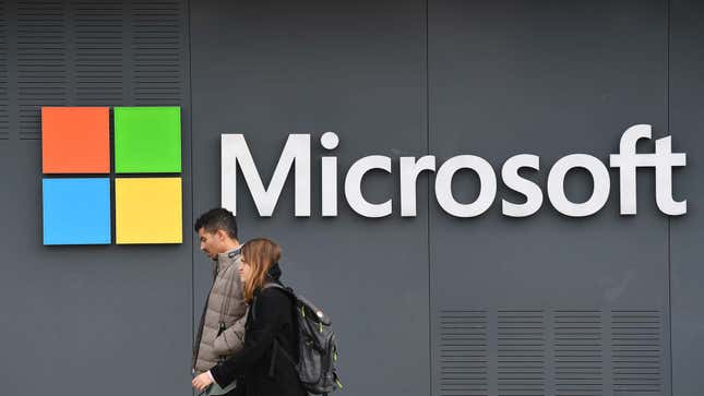A photo shows two people as they walk by a Microsoft logo on a grey wall.