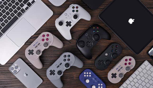 The entire collection of 8BitDo wireless controllers that now support Apple devices sitting on a wooden desk, surrounded by those Apple devices.