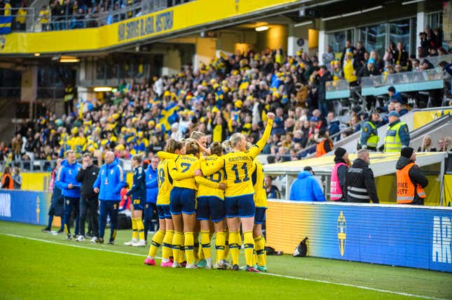 A group of women in matching yellow jerseys and blue shorts huddle and celebrate on a soccer pitch before a crowd.