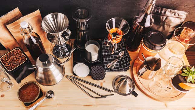 A counter filled with various coffee brewing tools and implements: a container of coffee beans, a bowl of ground beans, a goose-necked kettle, a pour-over brewer, several pitchers, and flavoring syrups