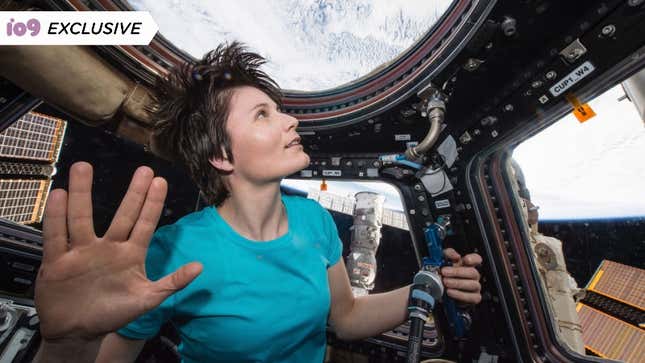 An astronaut holds up her hand in Star Trek's live long and prosper hand signal as she gazes out of her craft at the earth. She's wearing a plain blue t-shirt and her short hair is lifting upwards due to the lack of gravity.