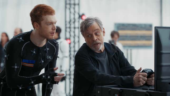 Mark Hamill sits at a TV with a controller while Cameron speaks to him in a black jumpsuit.