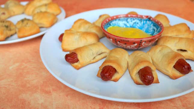 Mini hot dogs rolled in crescent dough on a plate with a small bowl of mustard in the middle.