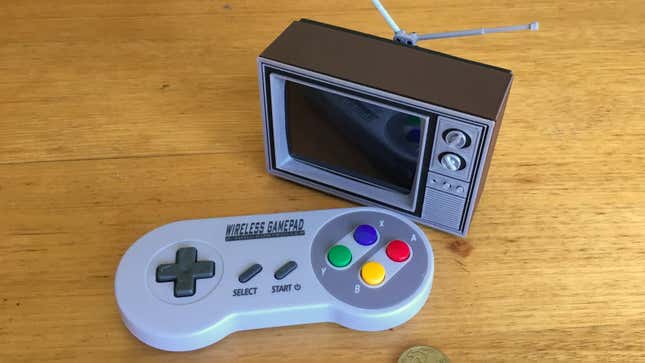 A tiny 3D-printed Toshiba TV featuring a modern LCD display sitting next to a wireless SNES-style gamepad on a wooden table.