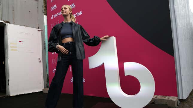 A woman in a black shirt, coat and pants holds up the TikTok logo to her right.