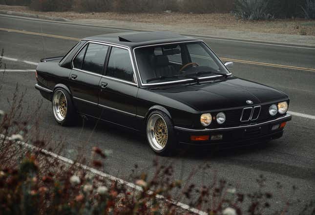 A modified black 1986 BMW 535is is parked on a California road with its headlights on.