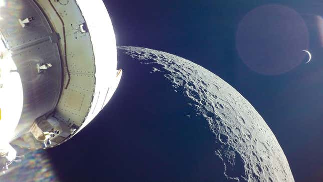 A photo taken from NASA's Orion spacecraft shows the cratered gray surface of the Moon, with Earth visible as a small crescent of light in the distance.