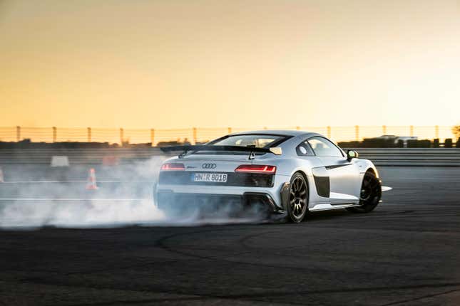 a grey 2023 audi r8 gt seen from the rear as it drifts around a curve on a race track in front of an orange sunset. tire smoke is pouring from the rear wheels