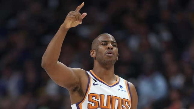 A Black man with close-cropped hair in a Phoenix Suns uniform, in front of a black background, holds up his hand to call a play during an NBA basketball game. 