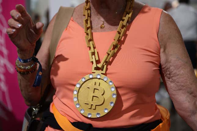 A person in a salmon-colored shirt wears a massive fake gold chain around their neck the the Bitcoin symbol at the end of it.