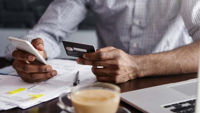 A man sitting at a desk covered in paperwork holds a credit card up next to his smartphone