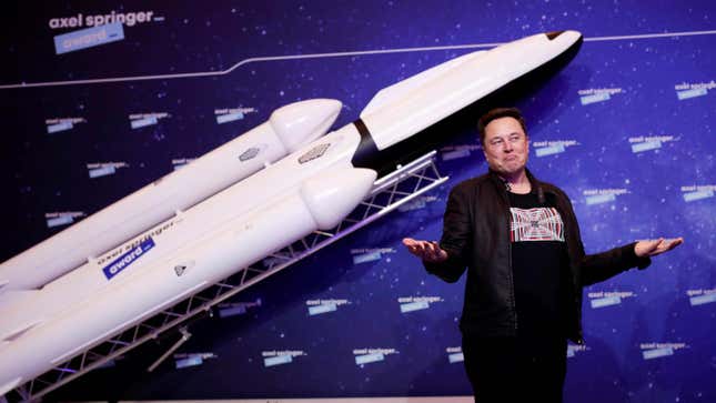 Elon Musk stands in front of a model rocket making a "so what" gesture with his hands.