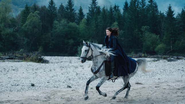 Moiraine rides swiftly on a white horse over a rocky beach, her blue cape flapping in the wind.