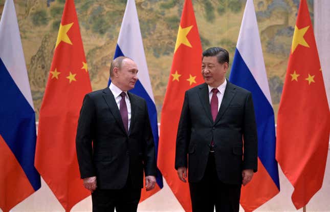 Vladimir Putin (left) and Xi Jinping (right) stand side-by-side in suits in front of alternating Russian and Chinese flags.