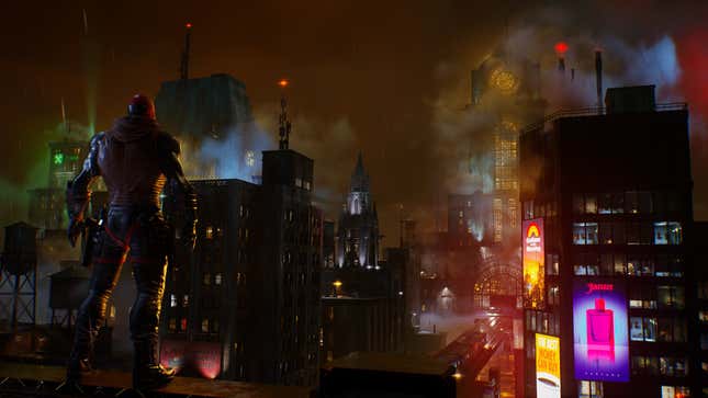 A man wearing a red helmet and armor stands on a dark rooftop overlooking Gotham City at night. 
