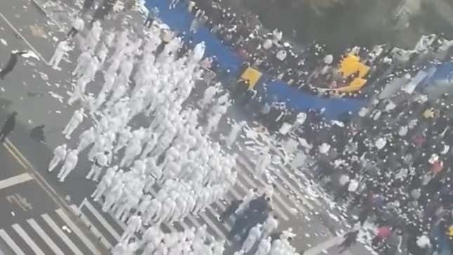 A hoard of white suited security officers stand opposite a large crowd of protesters on a dirty city street.