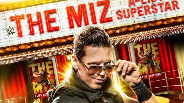 WWE superstar The Miz lowers his sunglasses and poses in front of a marquee and posters displaying his name. 