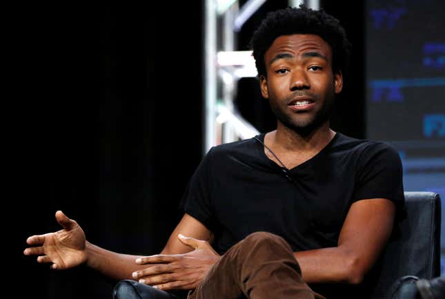 Donald Glover sits onstage in a black t-shirt explaining something to the audience. 