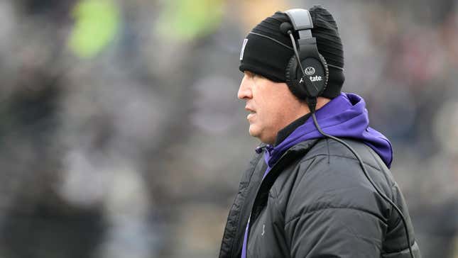A white man in a black puffer jacket, a black cap, a purple hoodie, and headphones is shown in profile on the sidelines of an NCAA football game. 