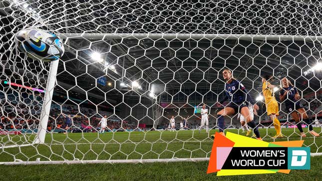 In a view from behind a soccer goal, a soccer ball looks as though it will burst through the net, as two women in matching blue soccer uniforms look on.