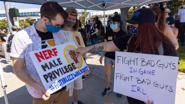Two protesting Activision Blizzard employees wearing face masks and holding white signs that say "Nerf Male Privilege" and "Fight Bad Guys In Game, Fight Bad Guys IRL."